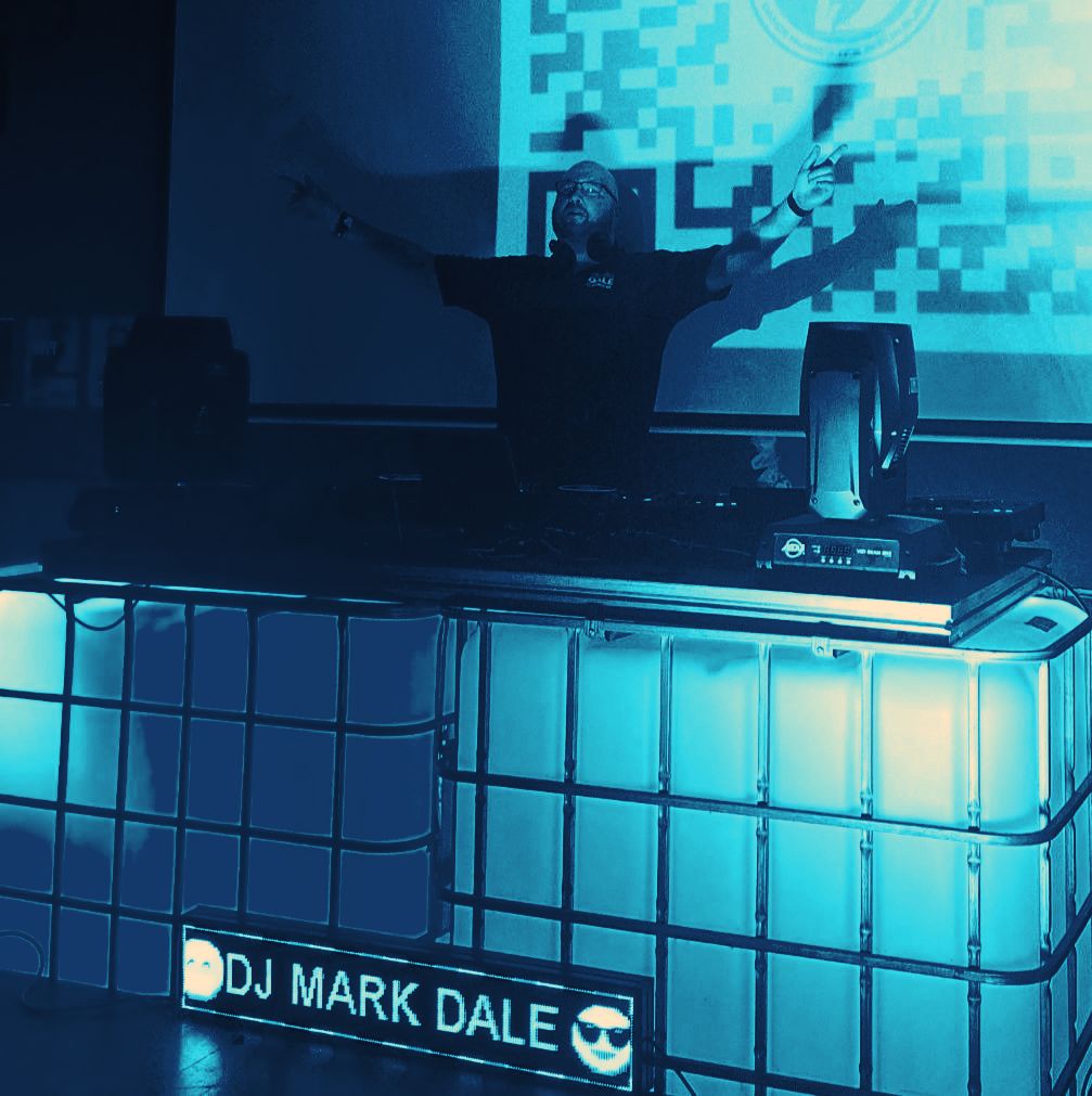 Mark Dale behind the decks with his arms outstretched, under pink club lights